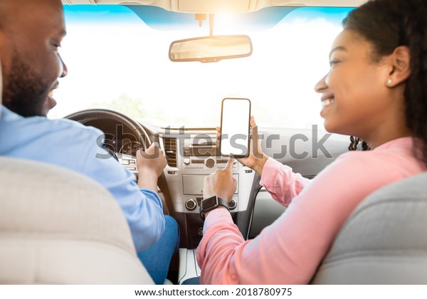 Look Here. Rear back view of happy African woman using\
cell, pointing at smart phone blank white screen inside a car,\
searching location via gps navigator application, man driving\
automobile, close up
