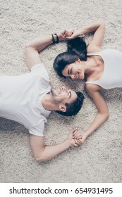 Look in my eyes forever. Dreamy couple of young beautiful lovers are lying on the beige cozy carpet indoors at home, holding each others hands. So sensual