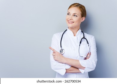 Look Over There! Confident Female Doctor In White Uniform Looking Away And Pointing While Standing Against Grey Background