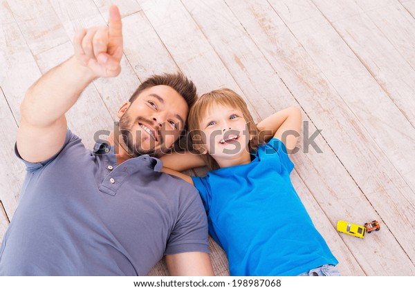 Look over there! Top view of happy father and son\
lying on the hardwood floor together while young man pointing away\
and smiling