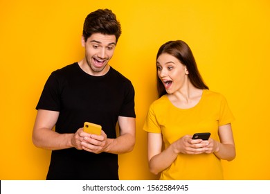 Look! Photo of two crazy students people guy lady holding telephones reading good news influencer blog open mouth wear casual black t-shirts isolated yellow color background