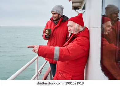 Look there. Senior man wearing protective jacket looking into the distance and pointing away with his finger while showing something to his colleague on the boat. Fisherman concept. Stock photo