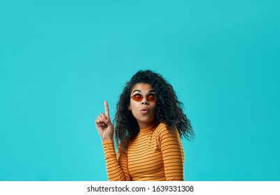 Look at this awesome nice advert! Smiling Afro American woman points thumb on copy space, recommends use it wisely, place your advertisement or promotion, wears orange sweater, has good mood