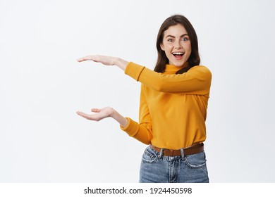 Look at this big thing. Excited smiling woman showing big object with hands on empty space, shaping box, sanding against white background.