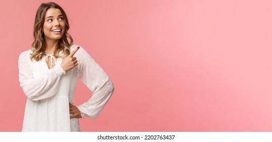 Look at this. Cheerful lovely blond european woman 20s in white cute dress, pointing at upper right corner with beaming smile, looking pleased, satisfied with good offer, pink background - Shutterstock ID 2202763437