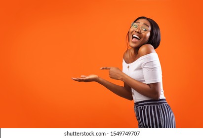 Look At This. Excited Afro Girl Pointing Finger At Something On Open Palm Standing Over Orange Studio Background. Free Space