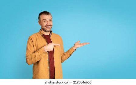 Look At This. Middle Aged Man Pointing Finger At His Hand Holding Invisible Object Advertising Your Product Standing Over Blue Studio Background, Smiling To Camera