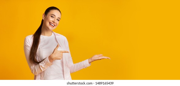 Look At This. Young Woman Pointing Finger Holding Something On Hand On Yellow Studio Background. Free Space, Panorama