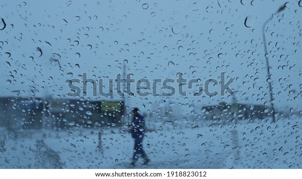 a look through\
glass in drops of wet snow at a pedestrian walking along the\
street, a blurry view of urban space in gray tones from a car\
window covered with a texture of\
drops