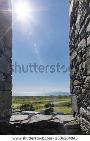 Look through large opening in wall of fortification Harlech Castle in Harlech, Gwynedd, Wales, UK built by Edward I during invasion of Wales with bright sun streaks over surrounding landscape