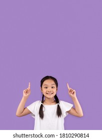 Look Up. Adorable Asian Kid Girl Pointing Fingers Upward At Copy Space Standing Over Purple Studio Background. Vertical