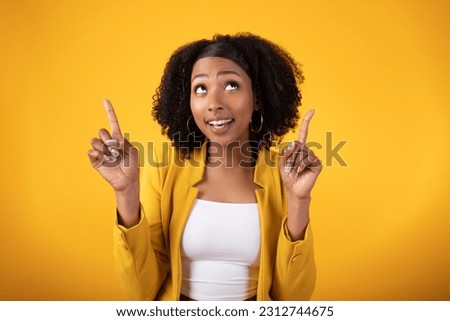 Look up. Excited black lady pointing fingers upward with both hands, looking and smiling, posing standing over yellow studio background. Check this offer