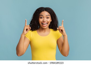 Look upward. Overjoyed african american woman pointing fingers up, looking and smiling to camera on blue background, studio shot. Black lady advertising something upward