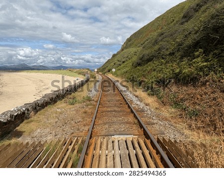 Looking along a train track at a level crossing on the British coast. The beach is on the left side of the track, with lush green hillside on the right. This level crossing and beach is in the town of Harlech, Wales. 