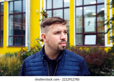 Looking Away Young Caucasian 34 Years Old Man In Blue Jacket With Goatee Beard On Yellow Facade Of College Modern Building. Young University Teacher And Scientist. Head Shot Man Lifestyle Portrait.