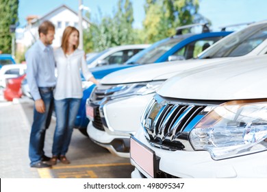 Looking For The Best Car Deal. Selective Focus On A Car Couple Choosing A Car At The Dealership Lot On The Background 