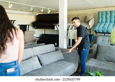 Looking To Buy New Furniture. Hispanic Couple Measuring A Sofa To See If It Will Fit In Their House