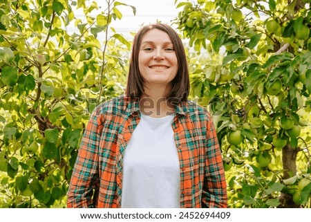Looking at camera front view stand in orchard of fruit trees young farmer woman smiling joyful. Agronomy adult girl is glad harvest rich. Family garden, family business, young business is successful.