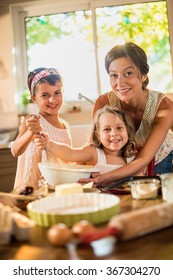 Looking At Camera, A Mom Is Cooking With Her Two Daughters Of Four And Seven Years Old. They Are Smiling, Wearing Casual Clothes. The Sisters Are Mixing The Ingredients In A Large Bowl With A Whisk.