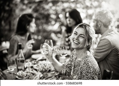 Looking At Camera, Portrait Of A Beautiful Middle Aged Blond Woman Sharing A Meal Friends On A Terrace Table In Summer, She Has A Glass Of Wine At Hand. Black And White