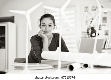 Looking at camera. Portrait of a beautiful woman architect, working on an ecological construction project in her office. There are models of wind turbines at the foreground. Black and white

