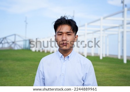 Looking at the camera portrait of a serious Asian guy. Young man standing outdoors. Confident people concentrated no smile. Young male person wearing blue shirt