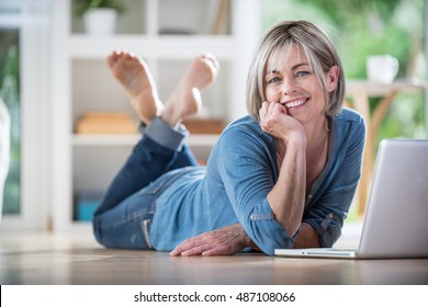 Looking At Camera. A Pretty Forty Year Old Woman With Gray Hair. She Is Lying  On Wooden Floor In The Living Room. She Consults Her Laptop. She Is Happy, She Smiles. She Is Wearing Jeans And A Shirt