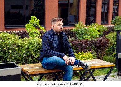 Looking At Camera Young Caucasian Businessman 34 Years Old Man With Goatee Beard Sits On Bench In Modern Landscaped Garden In Residential Complex. Full Length Lifestyle Portrait.