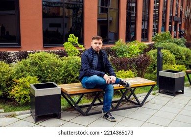 Looking At Camera Young Caucasian Businessman 34 Years Old Man With Goatee Beard Sits On Bench In Modern Landscaped Garden In Residential Complex. Full Length Lifestyle Portrait. Waiting