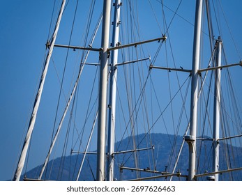 Looking up at the complex, intricate masts, spreaders, and rigging of a tall sailing ship, set against a clear blue sky and a distant mountain peak.