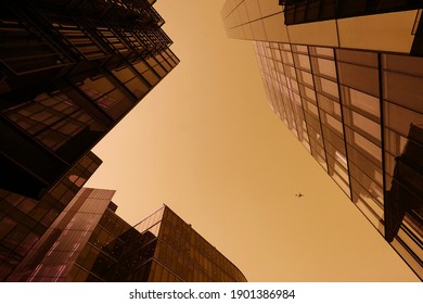 Looking directly up in the midle of glass corporate buildings, financial skyscrapers, a jet airplane crosses the business office towers in yellow golden orange. Modern, contemporary.low angle view 
