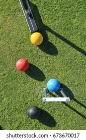 Looking down from above on a game of croquet with a croquet mallet about to strike the yellow ball with the blue ball in the jaws of the steel croquet hoop with the black and red balls nearby.