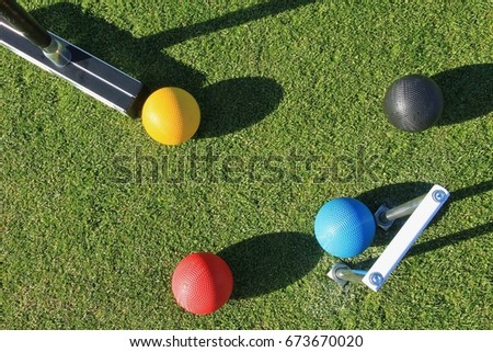 Looking down from above on a game of croquet with a croquet mallet about to strike the yellow ball with the blue ball in the jaws of the steel croquet hoop with the black and red balls nearby.