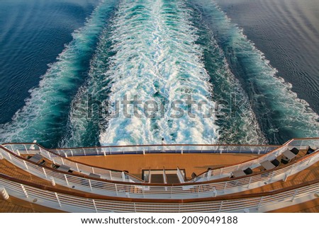 Looking down at the aft or back decks of a cruise ship and the wake or waves in the sea as it sails across the ocean. Open decks with tables and chairs and railings to view and relax. 