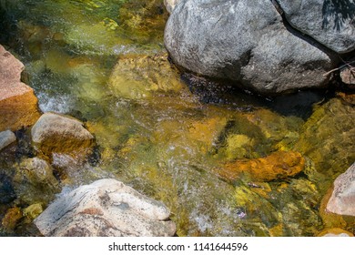 Looking down at a clear river flowing over stones, large granite boulders on each side.