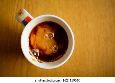 Looking Down At A Glass Mug Of Coffee, On A Light Wood Table, Just After Milk Has Been Poured Into It. This Creates A Milk Cloud Inside The Mug. Some Froth And Bubbles On Top. Copy Space To Add Text.