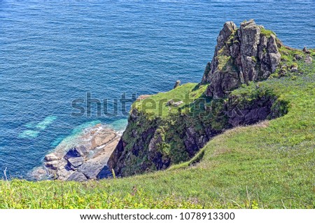 Looking down from a hilly plain on the Cornwall coast with grass and flowers towards a rock formation stepping down towards the Keltic Sea