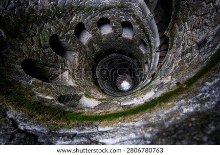 Looking down the Initiation Well spiral staircase in Quinta da Regaleira, mossy stone walls and arches descending into dark mysterious depth, Sintra, Portugal.