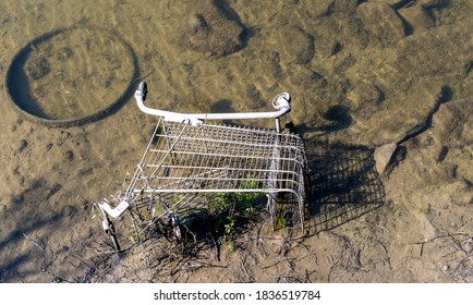 Looking down into a muddy river bed at a shiny, rust-free, stainless steel supermarket shopping trolley. The cart lies on its side partly submerged in silt with green weed growing through the mesh.