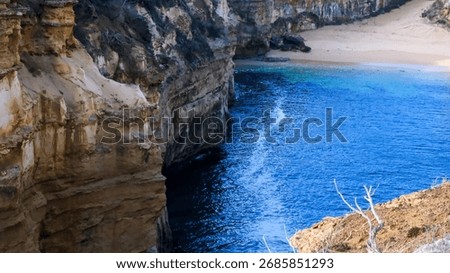 Looking down into the secluded cove of Loch Ard Gorge, revealing the deep blue water and a small patch of sandy beach between the sheer, dark cliffs.