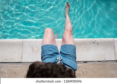 Looking Down At Man Sitting On The Edge Of The Pool With One Foot In The Water Wearing Blue Swimming Trunks. Fit Man Sits Poolside. Caucasian Man By An Outdoor Swimming Pool In The Summer.