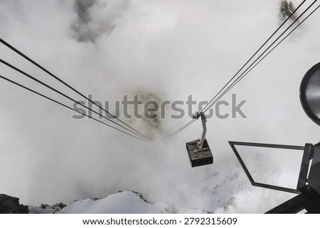 Looking down from a mountain platform at an aerial tramway cabin suspended on cables as it disappears into a dense blanket of fog over snowy terrain.