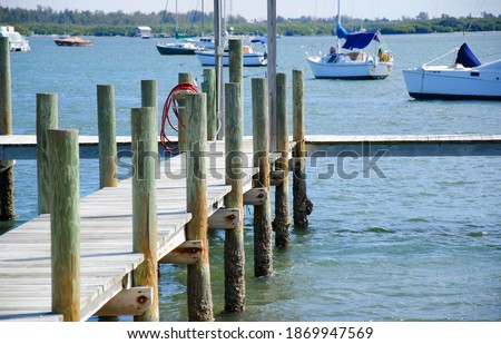 Looking down old wood T dock with barnacle covered posts, sunlit planks, rusty bolts, and bright red hose hanging on piling, with background of sailboats anchored in calm blue green waters.
