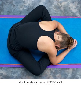 Looking down on an athletic brown haired woman doing yoga exercise Bound Angle Forward Bend pose on yoga mat in studio with mottled background.