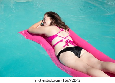 Looking Down On Cute White Woman Relaxing On A Pink Raft In A Backyard Swimming Pool Near A Diving Board On A Sunny Summer Day.  Girl Sunbathing On A Raft In A Swimming Pool. Getting Away From It All.