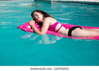 Looking Down On Cute White Woman Relaxing On A Pink Raft In A Backyard Swimming Pool Near A Diving Board On A Sunny Summer Day.  Girl Sunbathing On A Raft In A Swimming Pool. Eyes Closed Head On Arms.