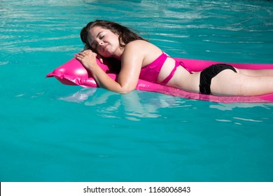 Looking Down On Cute White Woman Relaxing On A Pink Raft In A Backyard Swimming Pool Near A Diving Board On A Sunny Summer Day.  Girl Sunbathing On A Raft In A Swimming Pool. Eyes Closed Wet Hair.