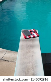 Looking Down On Folded Red And White Striped Beach Towel Folded On A Diving Board With A Book, Sunscreen, And Aviator Sunglasses On Top. Swimming Pool In The Backyard With Folded Towel On Diving Board