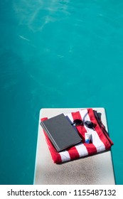 Looking Down On Folded Red And White Striped Beach Towel Folded On A Diving Board With A Book, Sunscreen, And Aviator Sunglasses On Top. Swimming Pool In The Backyard With Folded Towel And Novel.