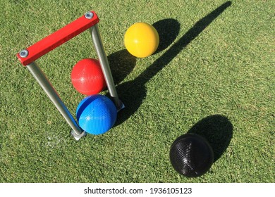 Looking down onto a set of red, blue, black and yellow croquet balls and a croquet hoop with a red cross bar on a green croquet lawn on a sunny day